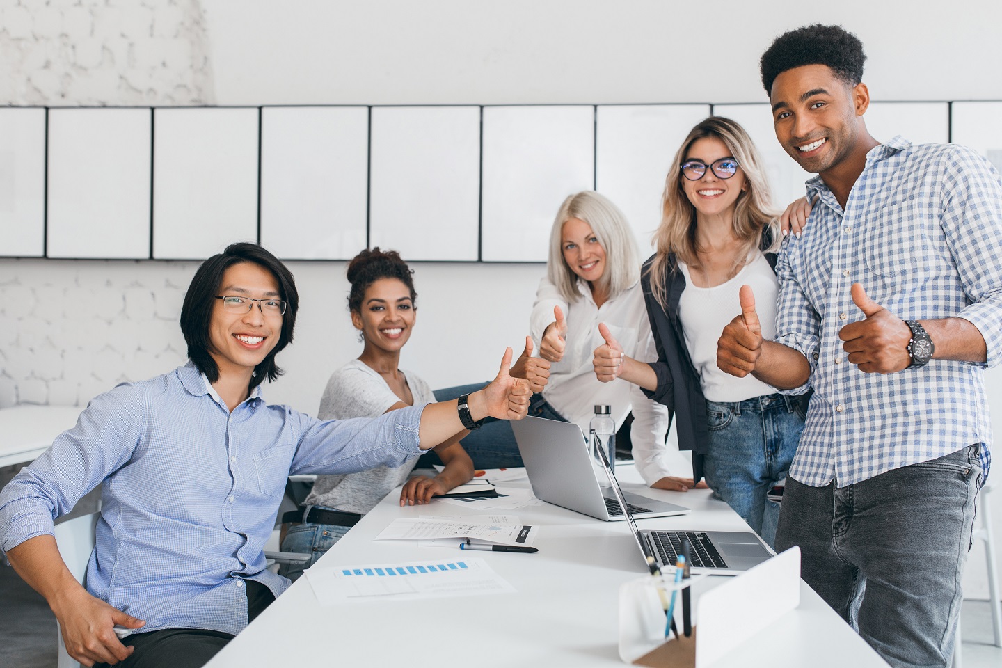 Blonde secretary sitting on table while office workers posing with thumbs up. Indoor portrait of happy asian manager in trendy shirt smiling in conference hall with foreign partners.